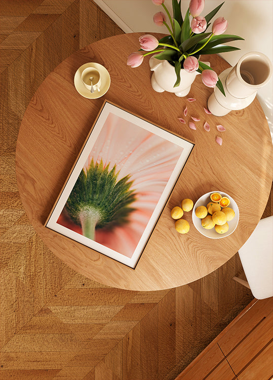 uma mesa de madeira com uma imagem emoldurada de uma flor, uma tigela de frutas e um vaso de flores rosa.