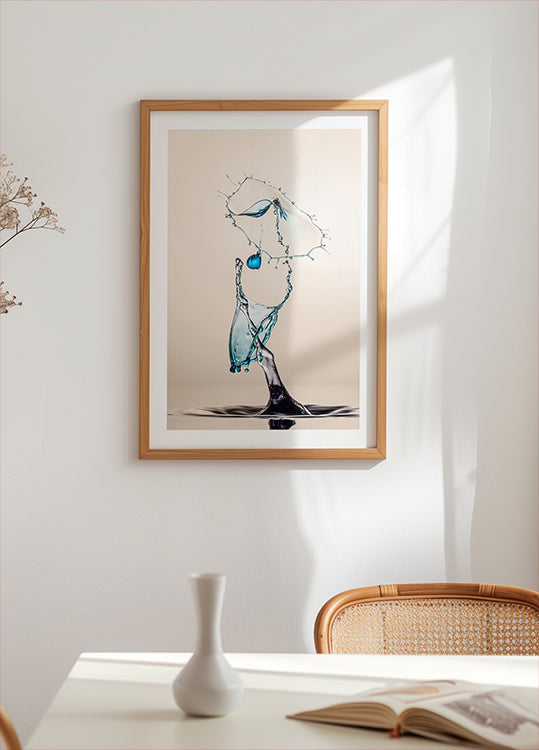 uma obra de arte emoldurada de uma gota de água suspensa no ar, com um esquema de cores azul e branco. A obra de arte está exposta em uma parede branca acima de uma mesa com um vaso branco e um livro.