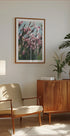 uma aconchegante sala de estar com armário de madeira, poltrona bege, quadro com flores cor de rosa e um vaso de planta.