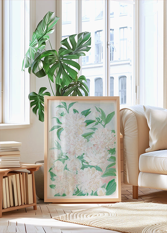 uma grande impressão emoldurada de flores brancas com folhas verdes, colocada sobre um piso de madeira em uma sala iluminada e arejada, com grandes janelas.