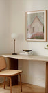 um espaço de trabalho moderno e minimalista com uma mesa de madeira, uma cadeira de tecido, uma luminária e uma pintura emoldurada de uma casa.